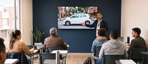 Instructor dicta una clase teórica frente a la pantalla con la imagen de un carro, ante cinco alumnos
